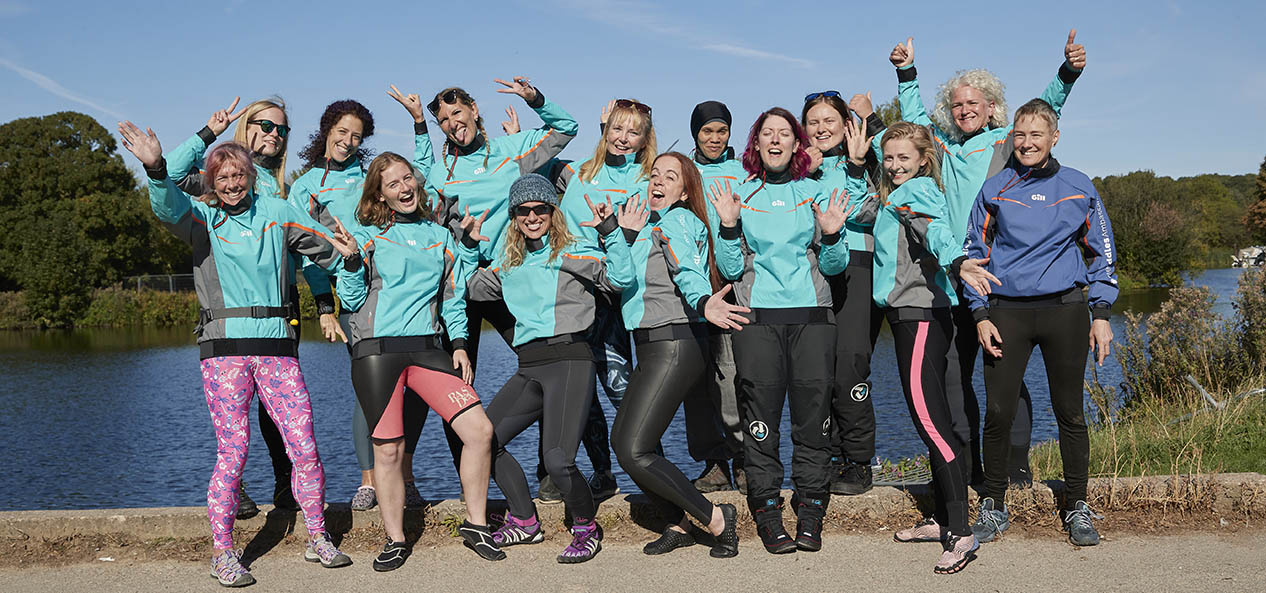 A group of women in canoeing kit pose for a photo beside a lake