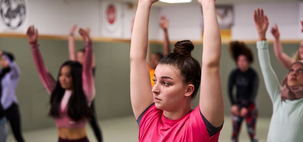 Young women in an exercise class in a sports hall raise their hands to the ceiling, they look very focused