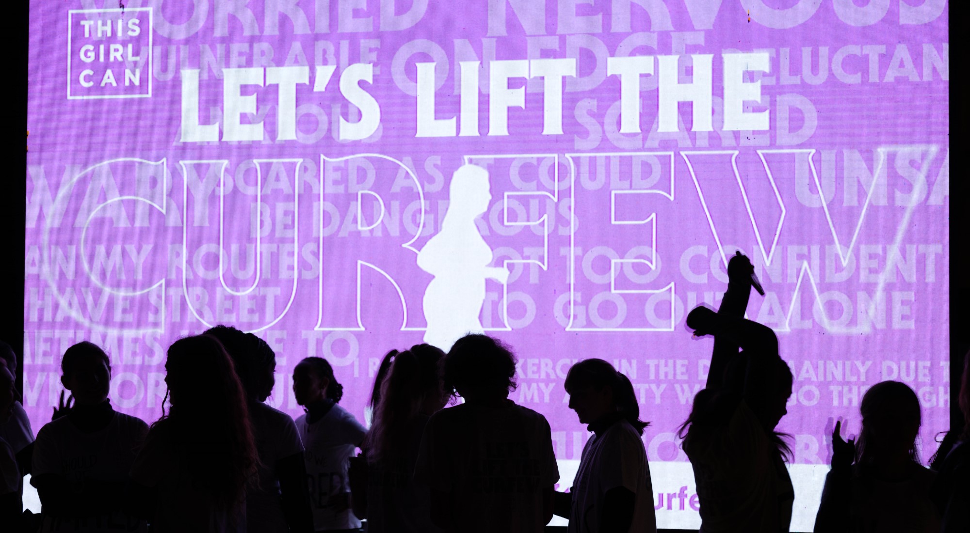 Silhouettes of women in front of an illuminated Let's Lift The Curfew sign.