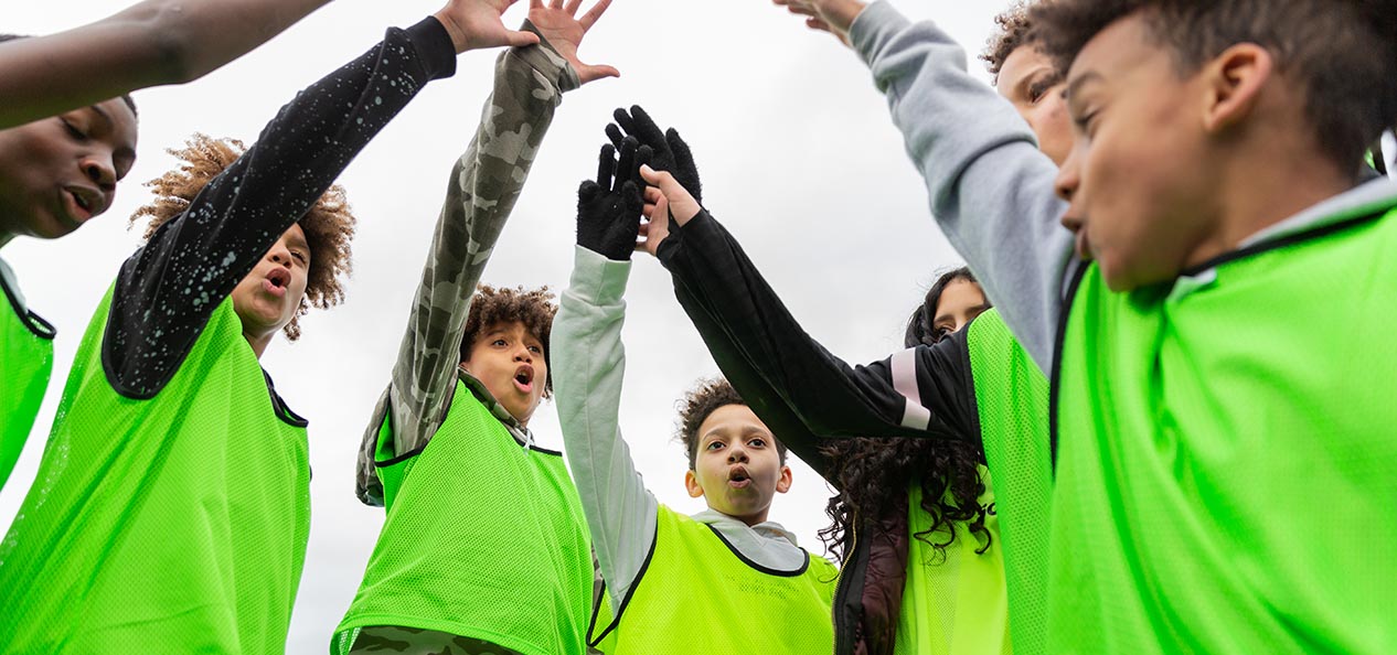 A group of seven kids in bright green vests celebrate by high-fiving each other outdoors.