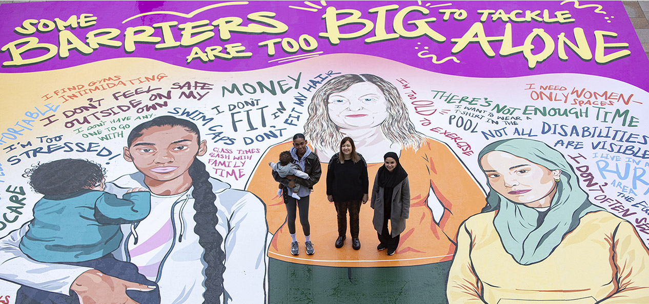 Three women, one holding their baby, stand on a mural painted on the ground, which carries the slogan 'some barriers are too big to tackle alone' - the mural also features a painting of the three women, standing in the pose they're stood in