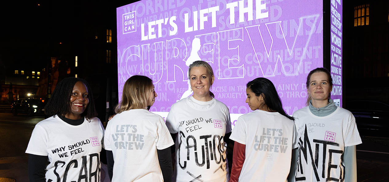 Five women stand in front of a lit-up bill board emblazoned with 'let's lift the curfew'. The women are wearing t-shirts printed with questions as to why they should feel 'cautious, scared, etc.' when getting active after dark