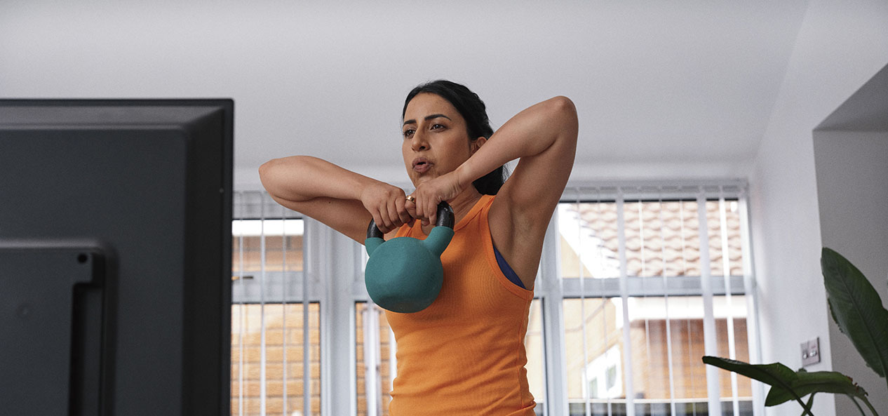 A woman lifts a kettle bell while doing a home workout in front of a TV at home