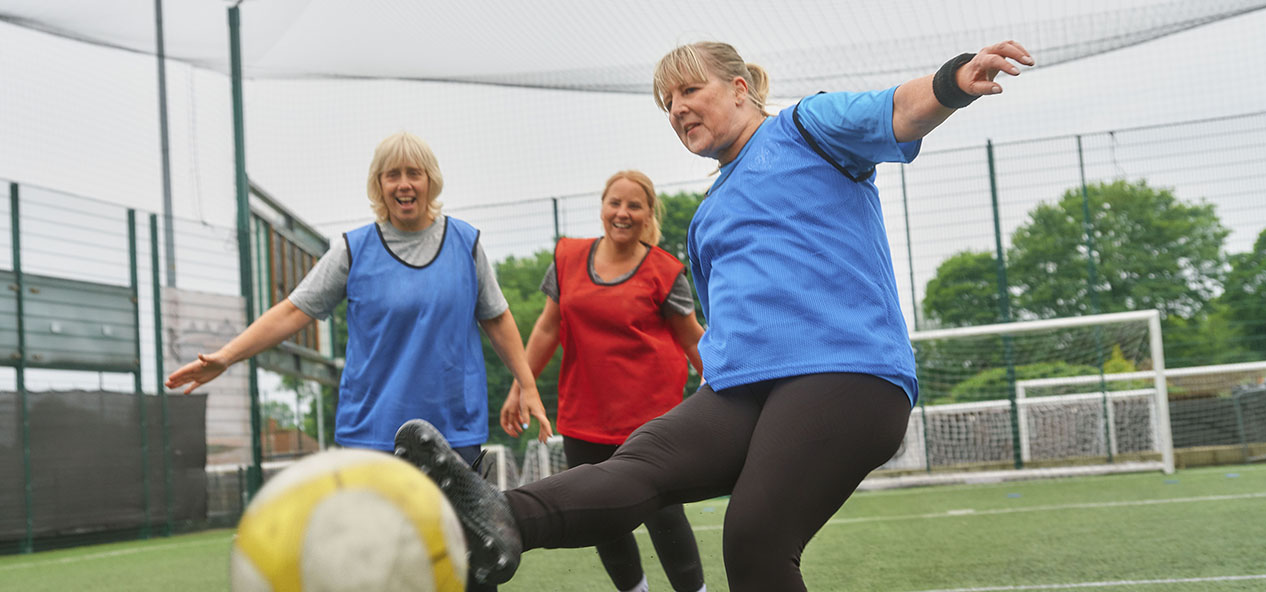 One woman, kicks a ball in a game of walking football, while two women - one teammate and one opposing player - watch on