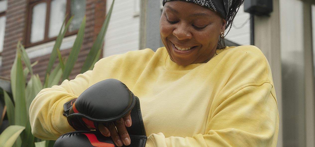 A woman in a yellow sweatshirt, does up boxing training gloves ahead of a home workout in her garden