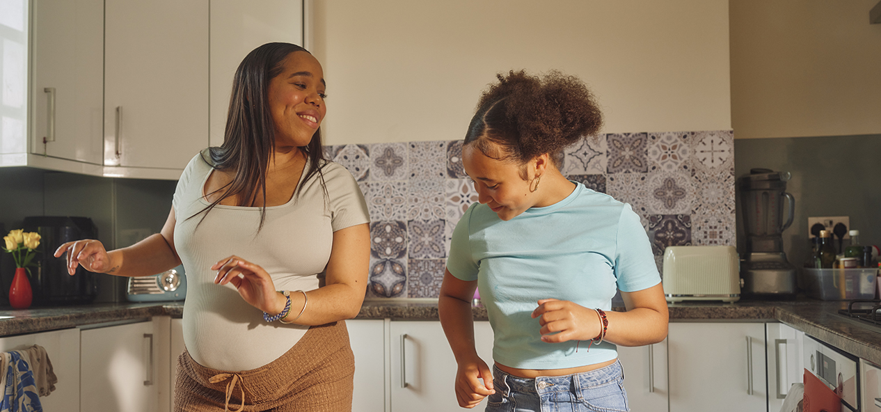 Two women, the one on the left being pregnant, dance in a kitchen