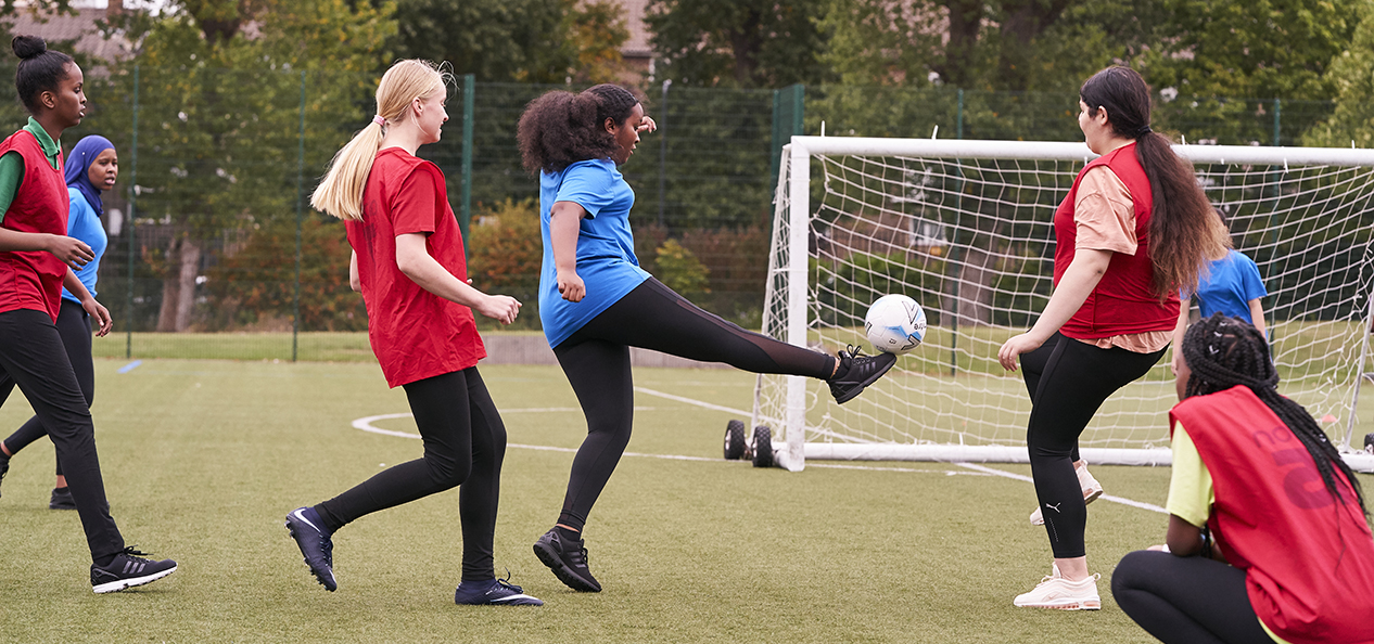 A group of White and Black girl, some of them wearing a headscarf, play football outdoors.