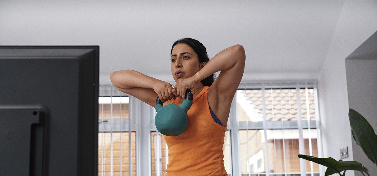 A woman lifts a kettle bell while doing a home workout in front of a TV at home