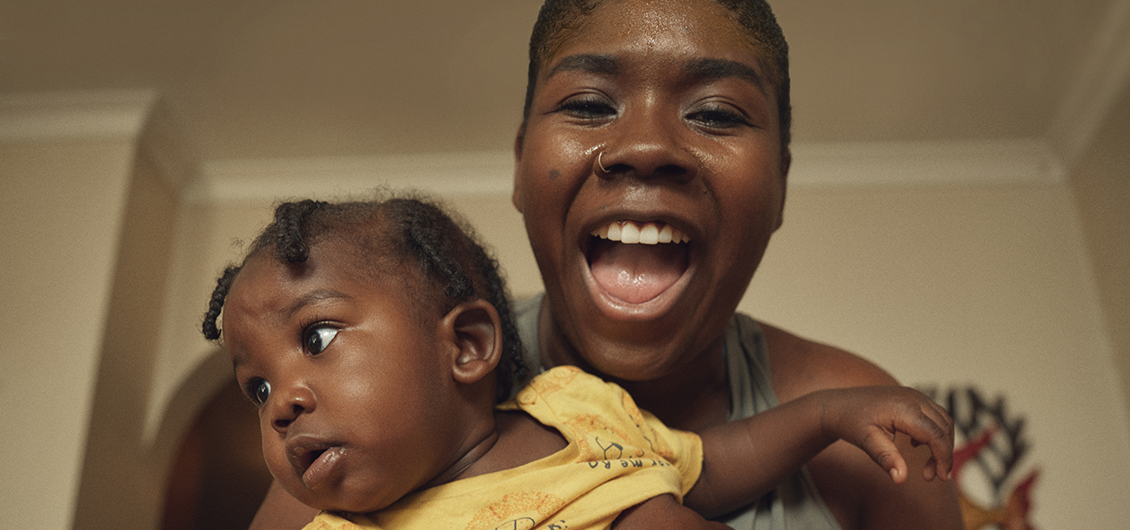 A black woman smiles at the camera, holding her baby in her arms