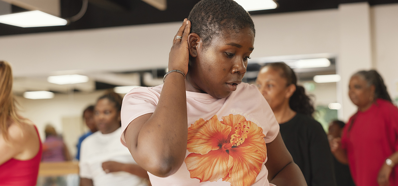 A black woman dances in front of others in a class, she has one hand on her head