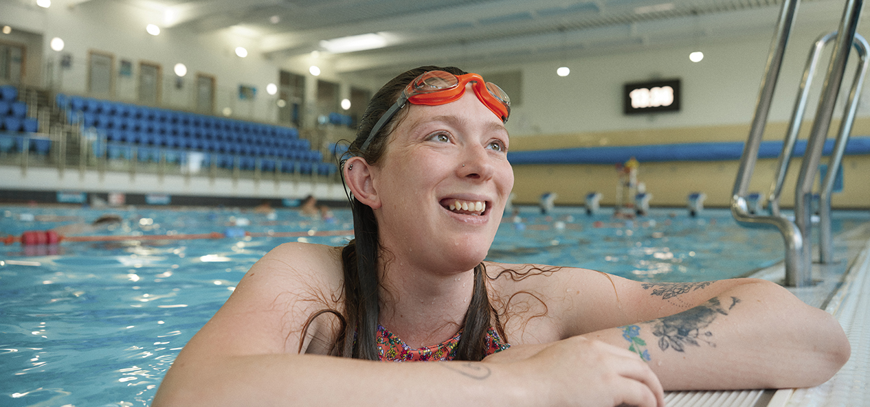 A pregnant woman wearing goggles leans on the edge of a swimming pool