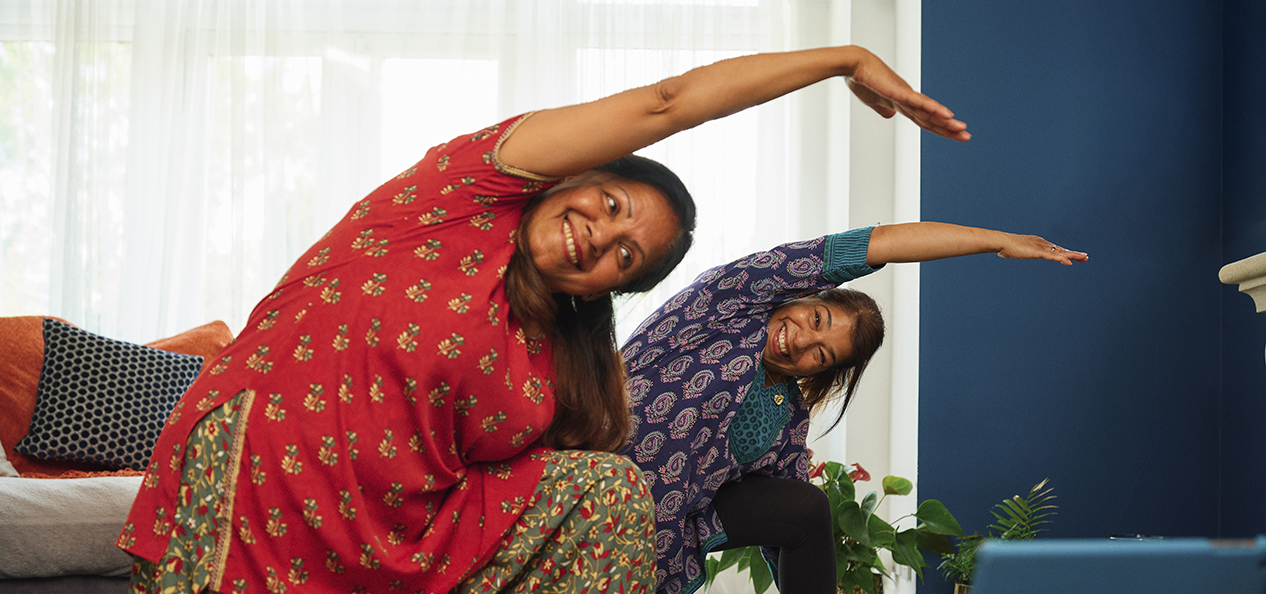 Two South Asian women perform yoga at home. Both face the camera, with one behind the other, both on one knee and bending to the side, with arm outstretched, over the other knee