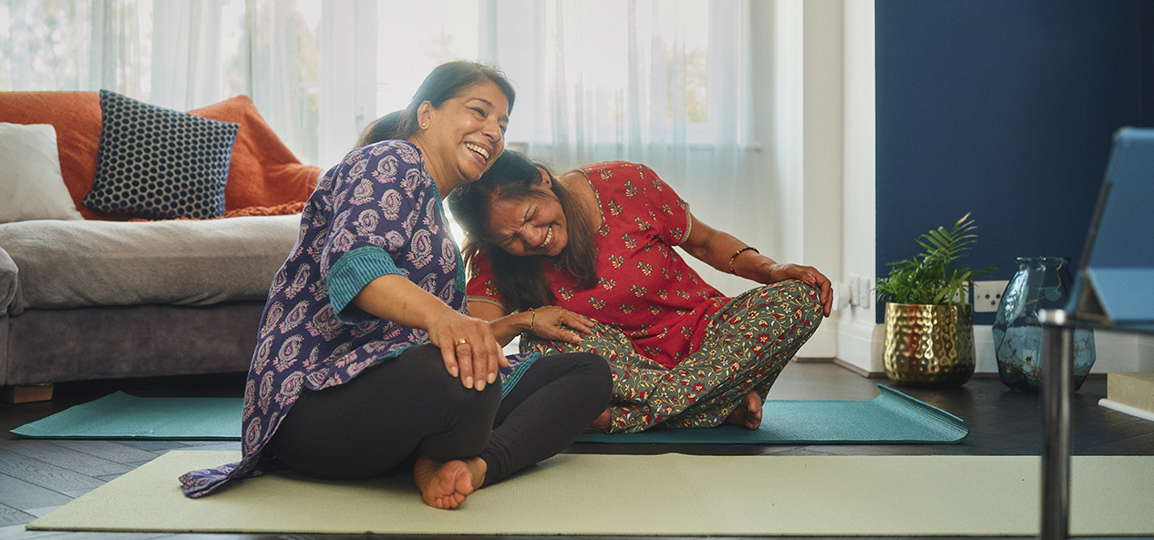 Two South Asian women sit crossed legged on yoga mats at home, both laughing and resting their heads on each other's