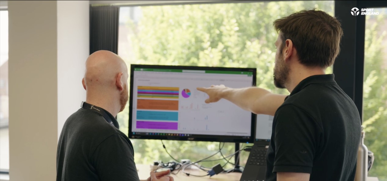 Two men stand looking at charts on a computer monitor, with one pointing at the screen.