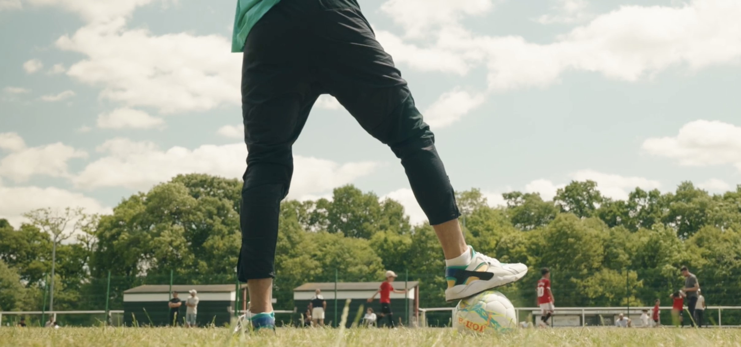 Close-up of a foot stood on top a football in the foreground of an amateur match, on a bright summer's day.