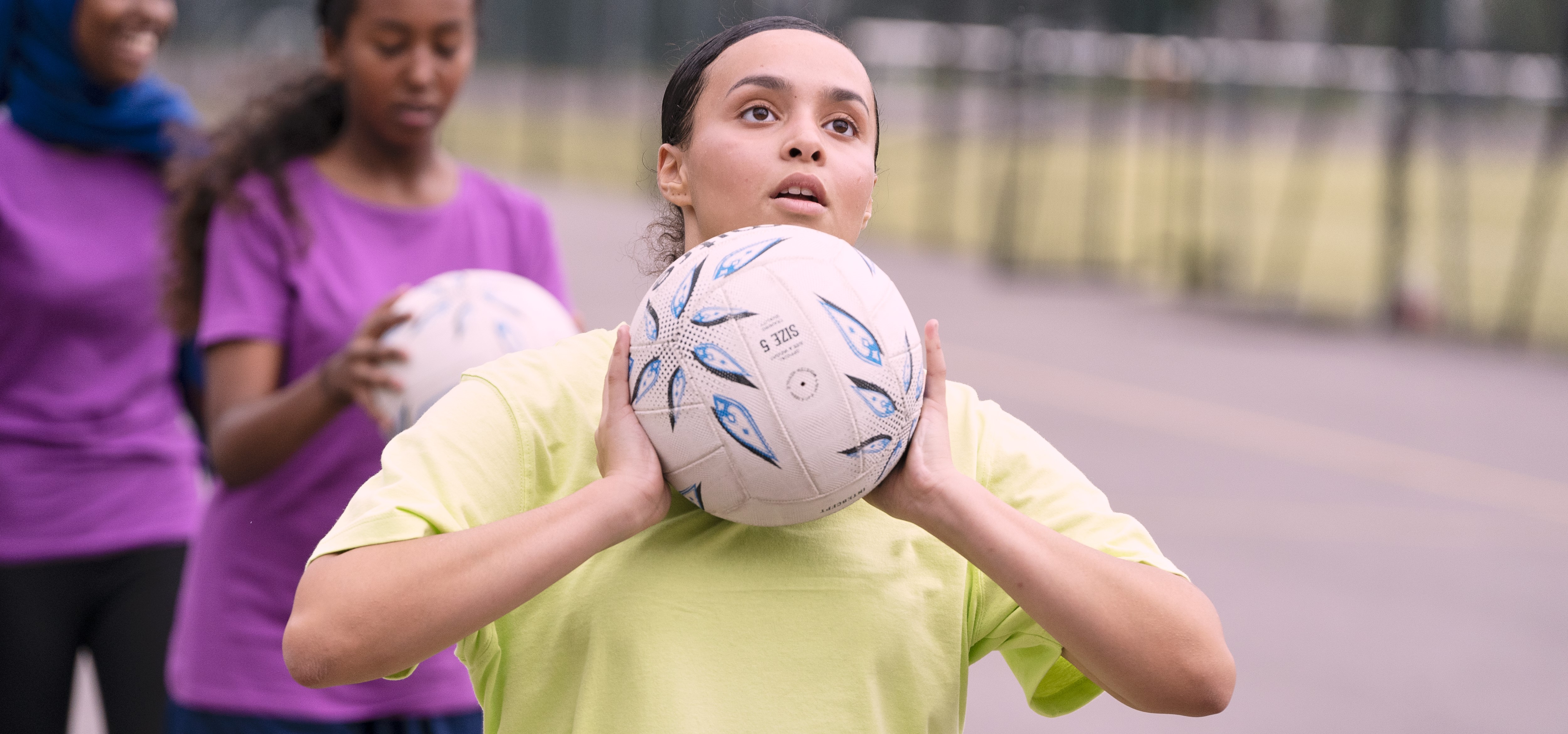 A girl aims to shoot a netball on an outdoor court, with two other girls queuing behind her to do the same.
