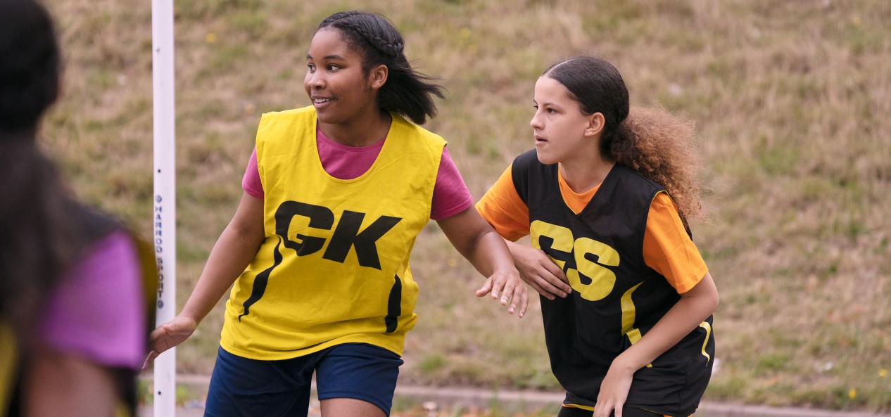 Two girls compete in a game of netball outdoors, wearing GK and GS vests.