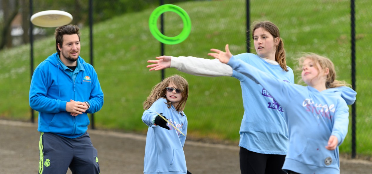 A volunteer watches as three girls throw frisbees on a playground.