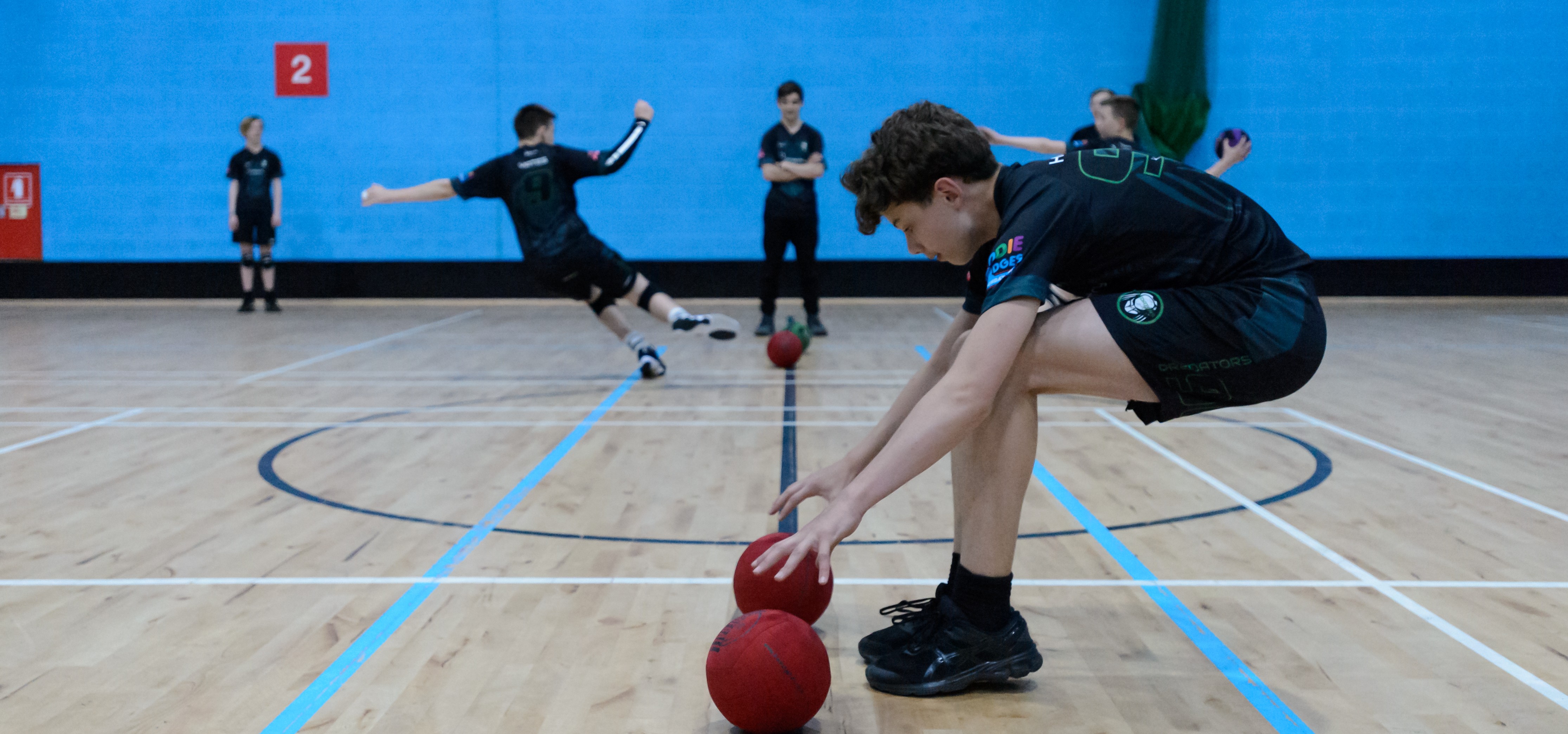 A boy bends down to pick up two dodgeballs from a sports ball hall, while other team-mates play in the background.