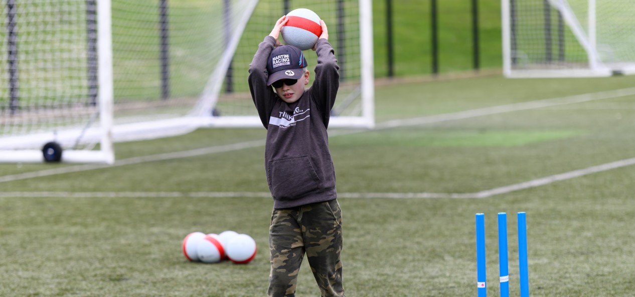 A boy wearing a baseball cap and dark glasses holds a ball above his head on an outdoor artificial football pitch.