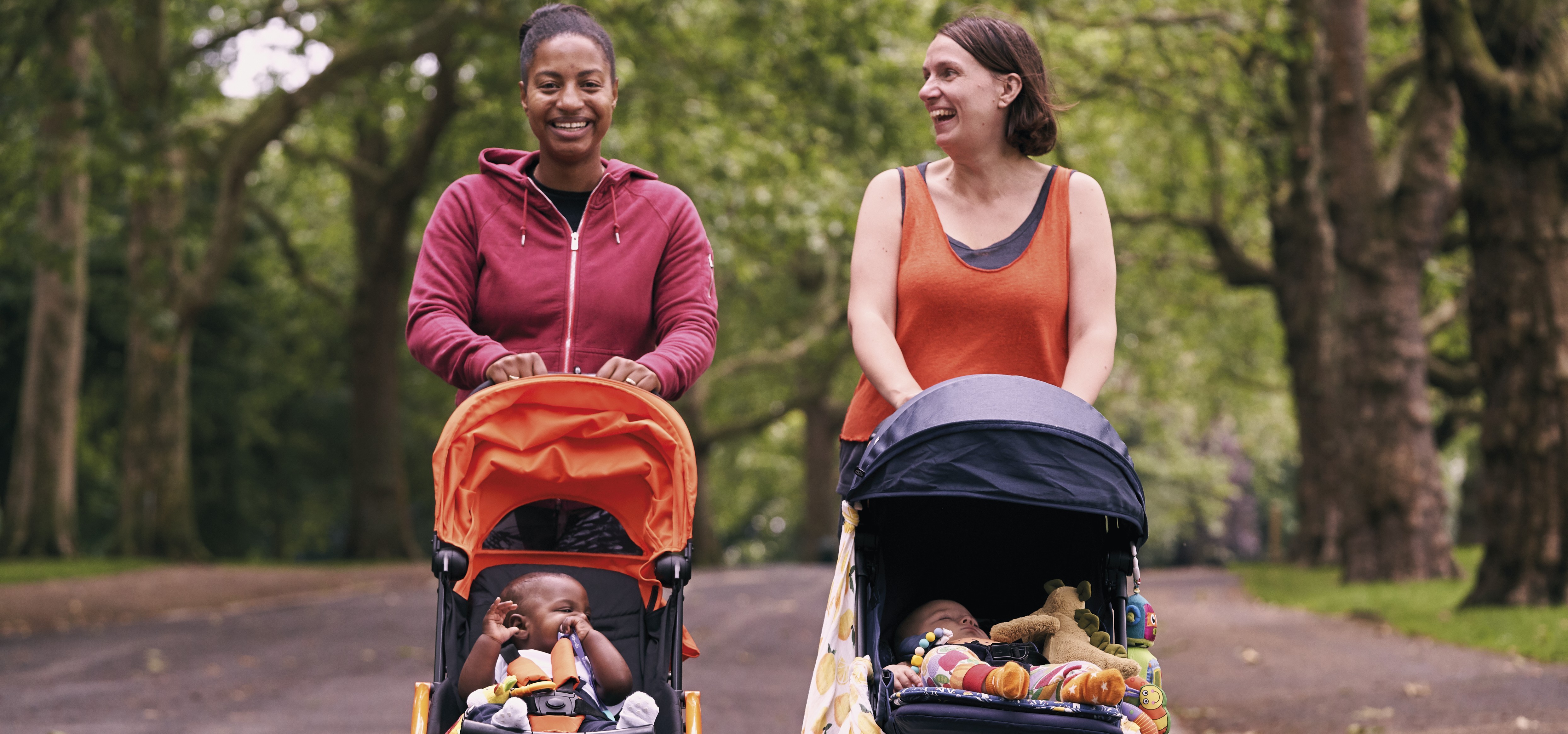 Two women in activewear walk side-by-side pushing prams with babies.