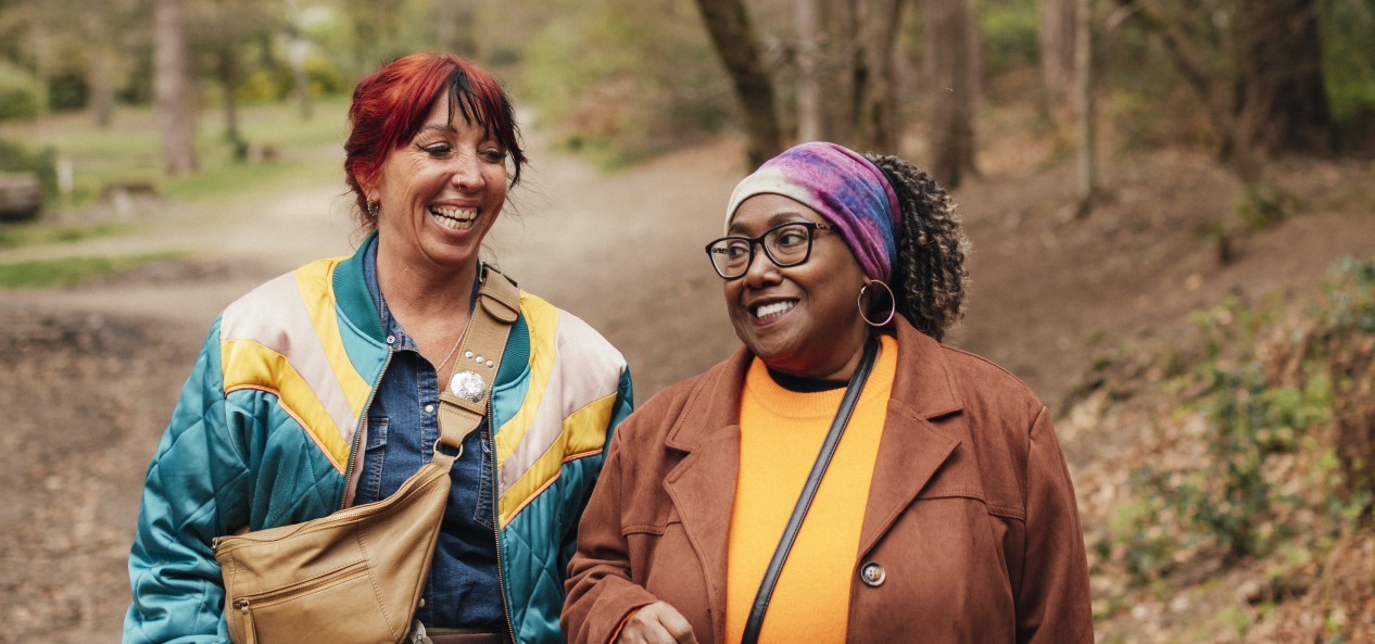 Two women link arms as they walk on a path in a forest.