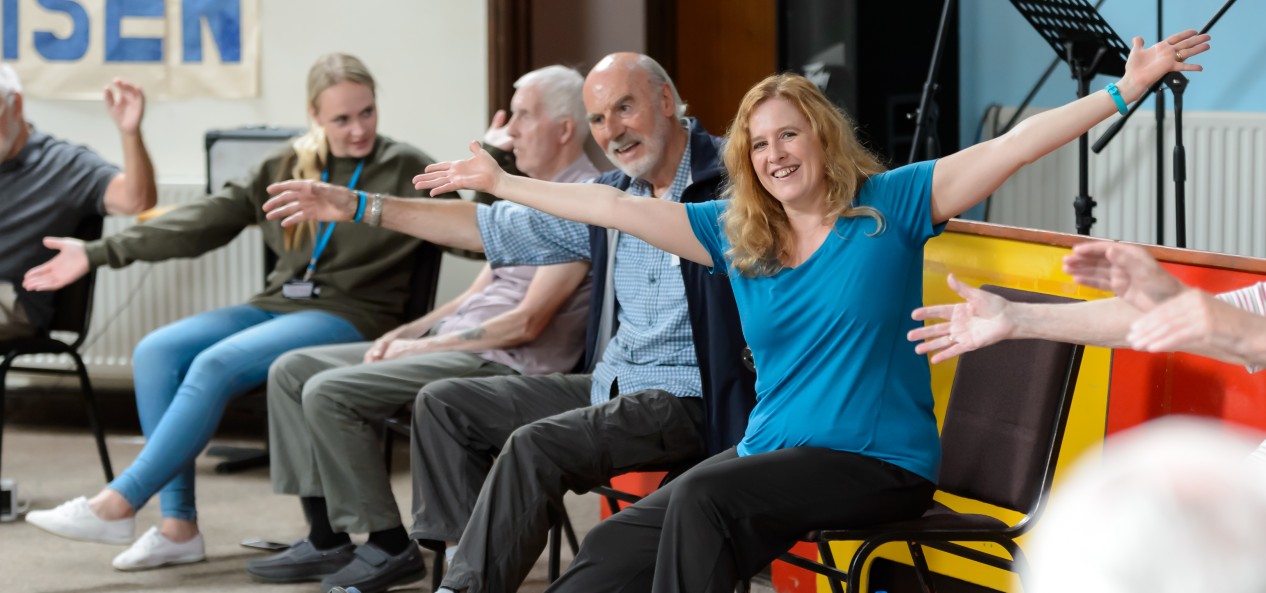 A woman stretches her arms alongside a row of older adults doing seated exercises in a hall.