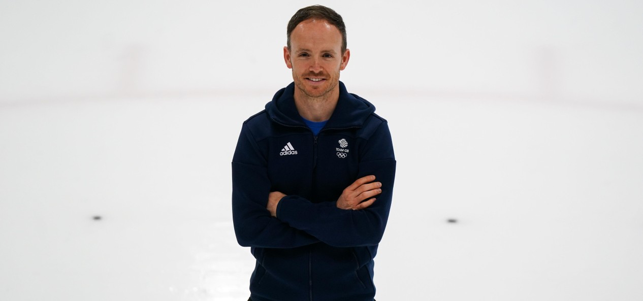 Team GB speed skating coach Richard Shoebridge smiles at the camera on the ice, with his arms folded.