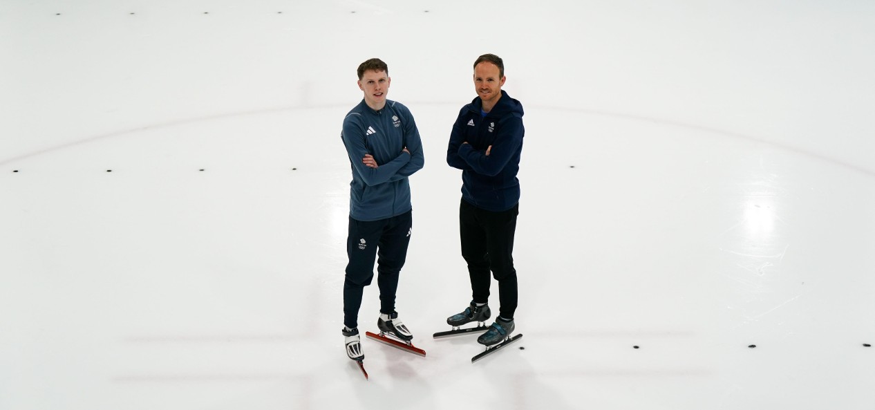 Team GB speed skater Niall Treacy and coach Richard Shoebridge smile at the camera on the ice, with their arms folded.