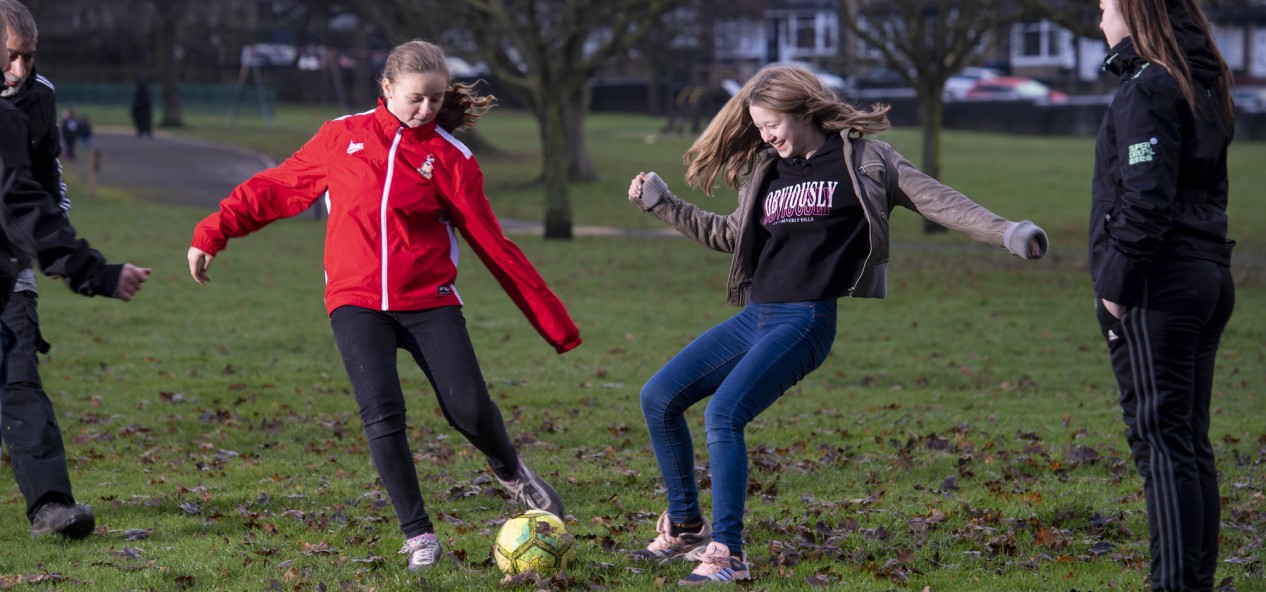 Two girls compete for a football during a casual game on an autumnal-looking park, while two fellow players look on.