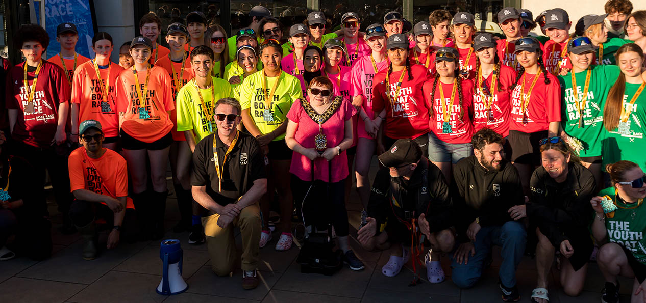 A group of young people with different colour t-shirts with "The Youth Boat Race" written on them pose with their medals on a sunny day.