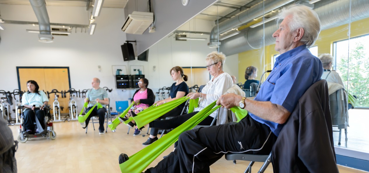 Several older adults use stretch their legs using resistance bands during a seated exercise class in a community hall.