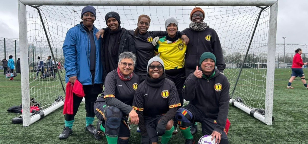 A group of eight women, all women of colour, standing and kneeling in front of a goal on a grassy football pitch. They're smiling at the camera.