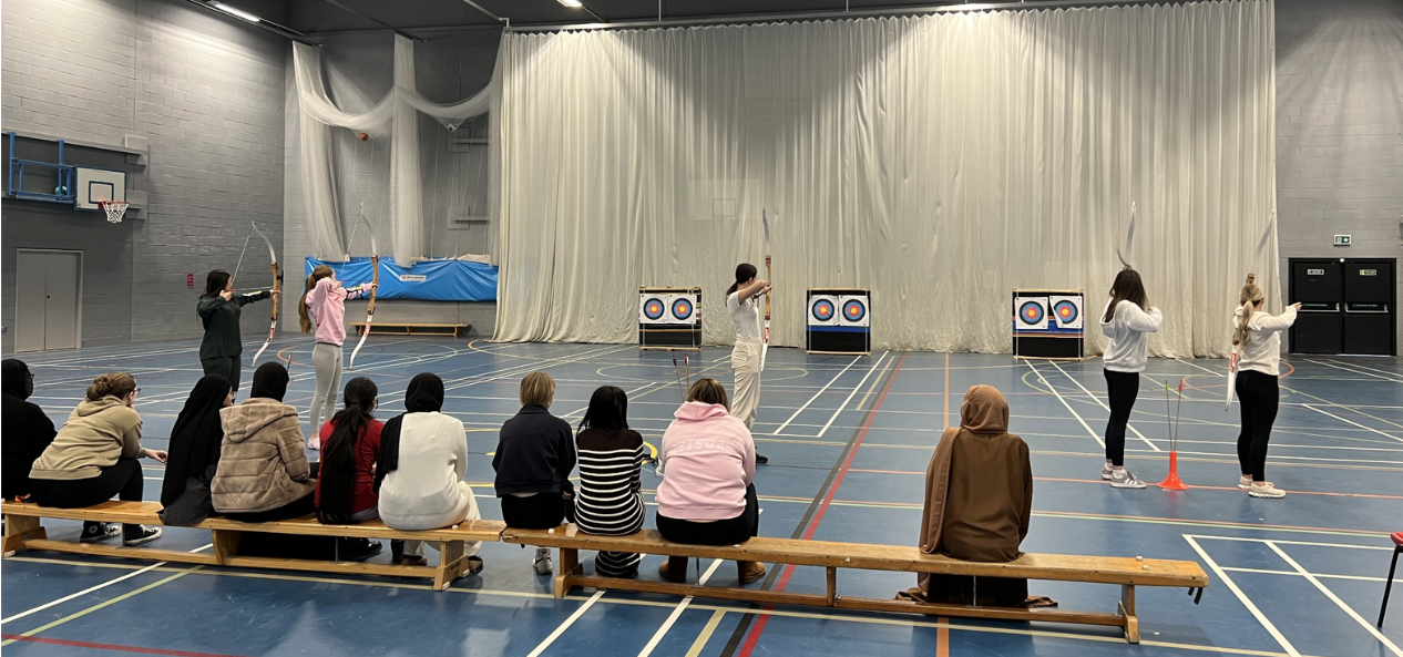 Girls sitting on bench in sports hall watching their peers taking part in archery