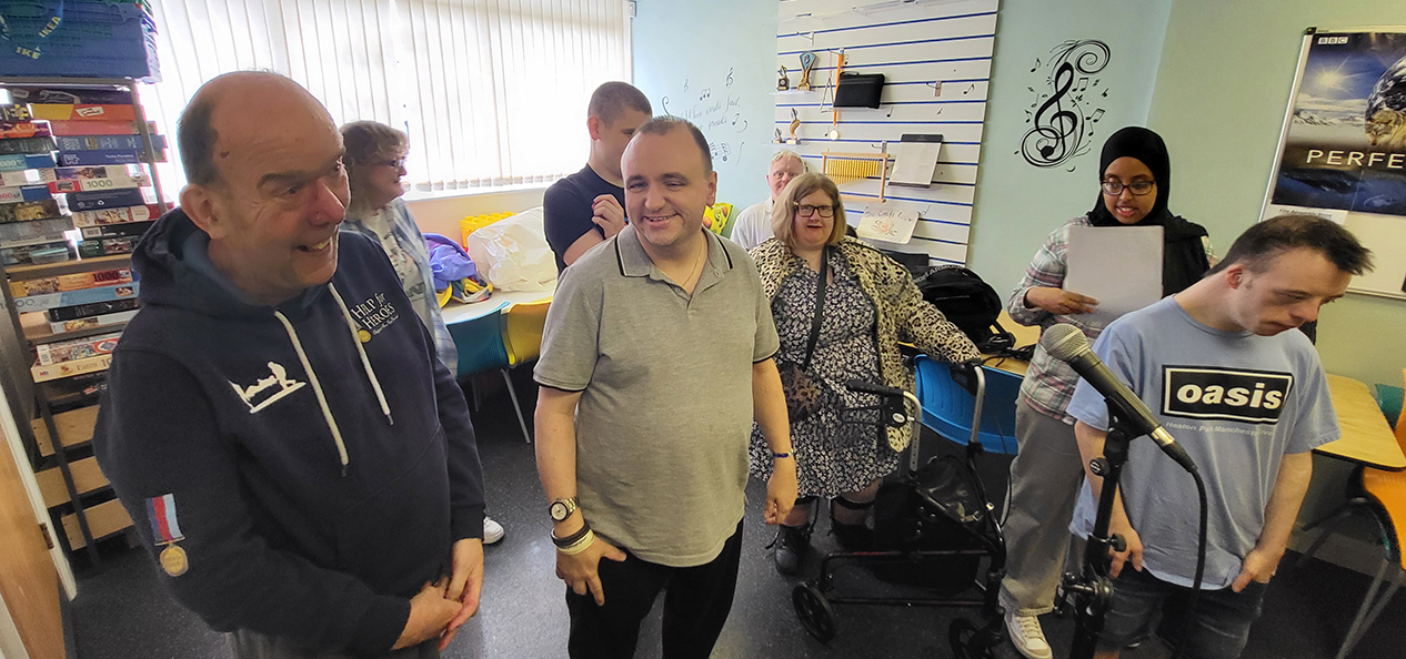 A group of adults with learning disabilities smile in front of some microphones inside a music room.