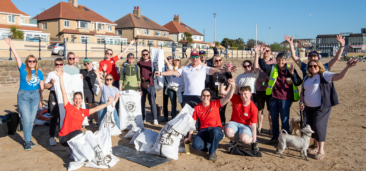 Paralympic champion Ellie Simmonds poses with a large group of smiling volunteers on a sandy beach on a sunny day, holding litter-picking bags and raising their hands. Several filled rubbish sacks are placed in the foreground, with a promenade, houses and a clear blue sky in the background. 