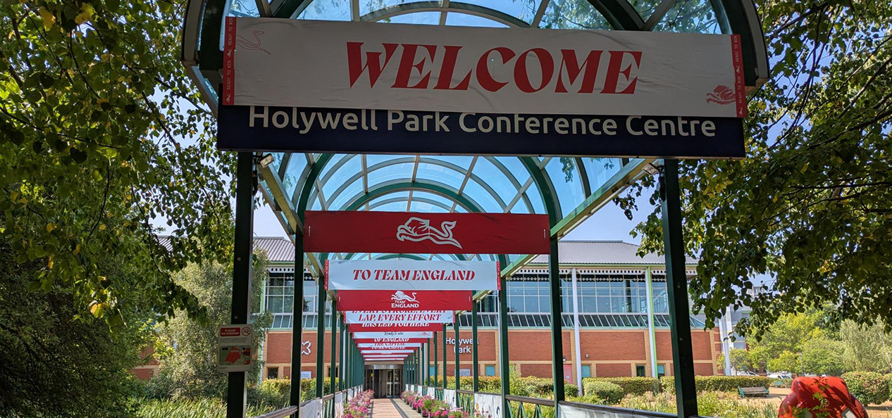 A view of the entrance of the Holywell Park Conference Centre, which is surrounded by trees and has a sign with "Welcome" in red letters on top of the name of the venue.