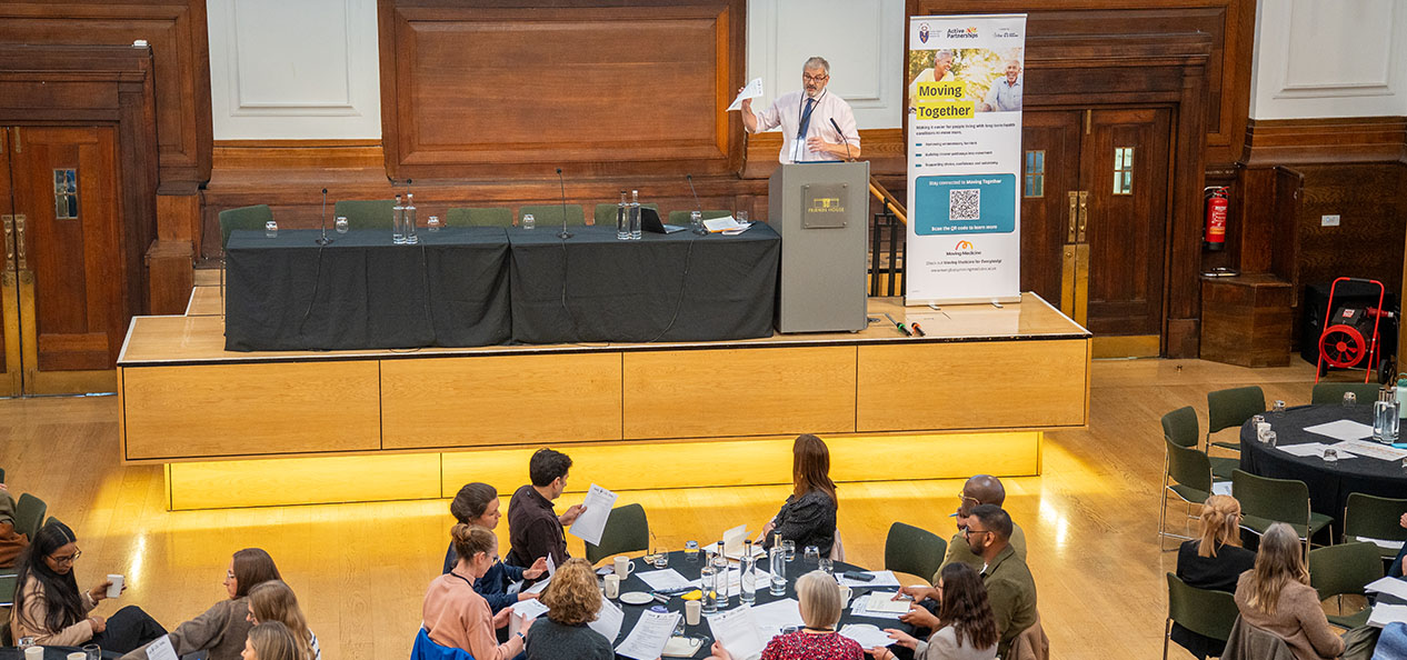 A man gives a presentation from a lectern to an audience seating around round tables in an indoors venue where there's also a Moving Together banner. 