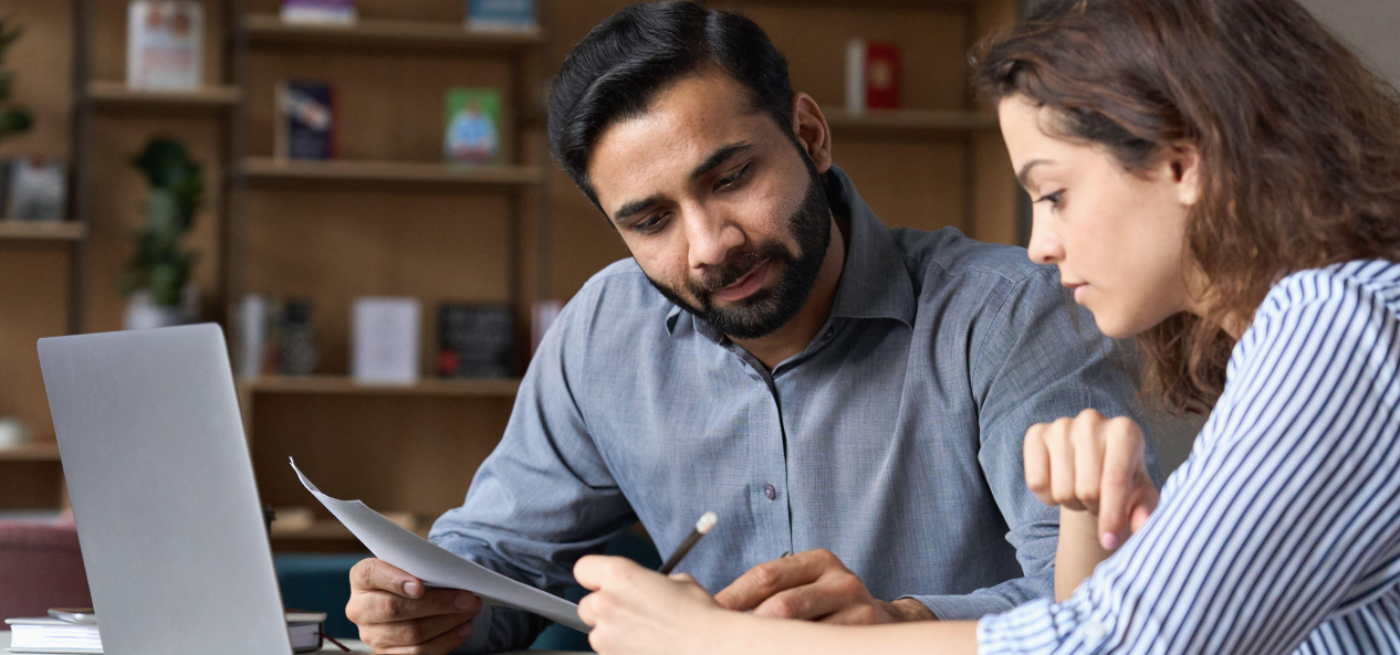 A man and a woman sit together looking at a piece of paper in front of a laptop in a work environment.