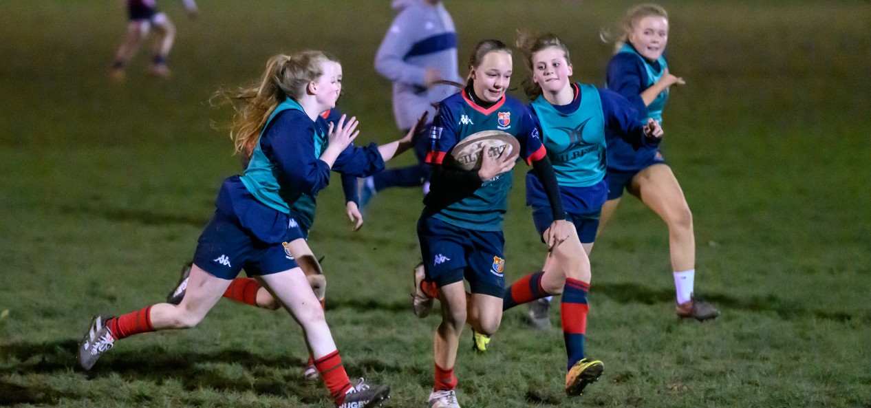 A young female rugby player is pursued by girls on the opposing team as she runs with the ball during a training game at night.