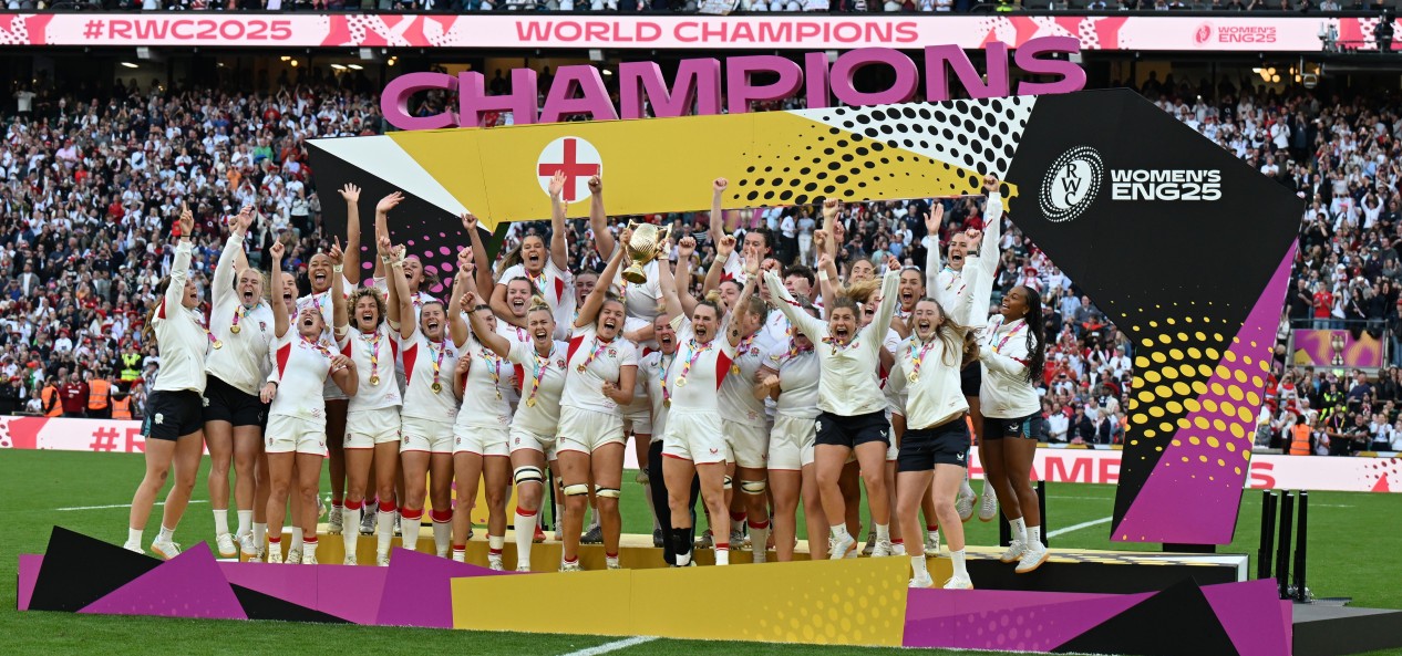 Zoe Aldcroft of England lifts the Women's World Cup 2025 trophy on the pitch at the Allianz Stadium in Twickenham, while her team-mates celebrate on a stage bearing the word 'Champions'.