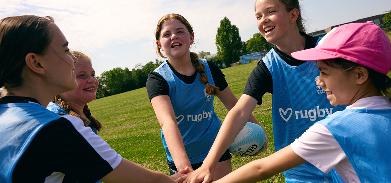 A group of girls put their hands together in front of them to show unity during an outdoors rugby session on a sunny day. 