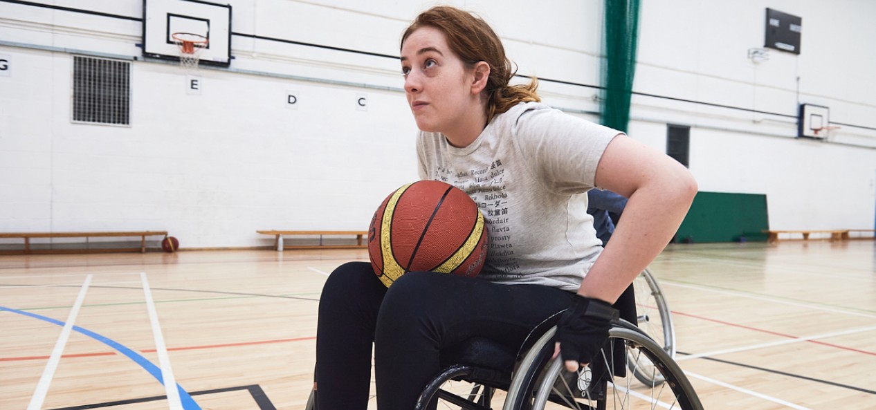 A female wheelchair basketball player advances with the ball on an indoor court.