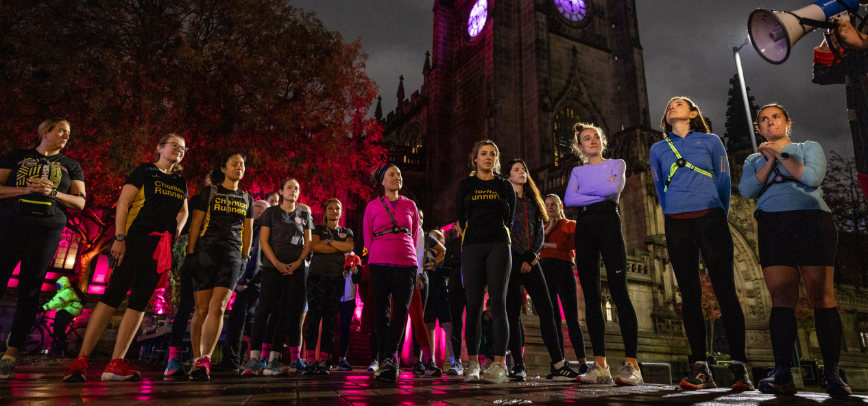 A group of women runners stand in a semi-circle under a clock tower at night, while someone to the side talks on a loudspeaker.