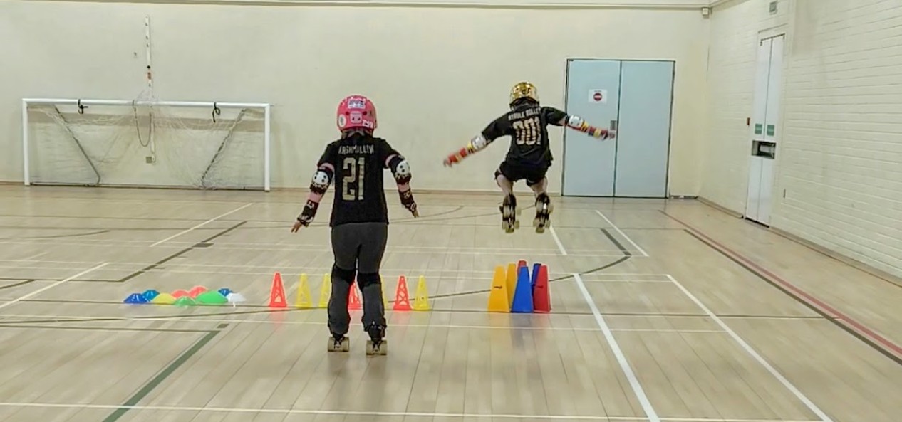 Two young people wearing helmets take part in a roller derby session, jumping over cones in a sports hall.