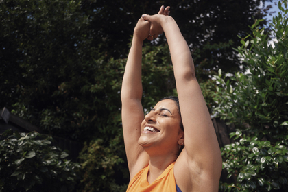 A woman closes her eyes as the reaches both arms above her head, clasping her hands together, smiling and tilting her head backwards while outside on a sunny day