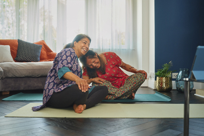 Two South Asian women sit crossed legged on yoga mats at home, both laughing and resting their heads on each other's
