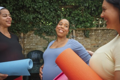 Three pregnant women stand talking outside while holding yoga mats.