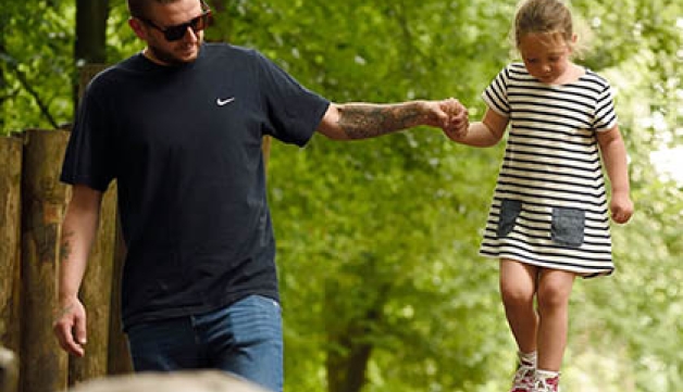 A father holding his daughter's hand as she walks along a tree trunk in a forest.