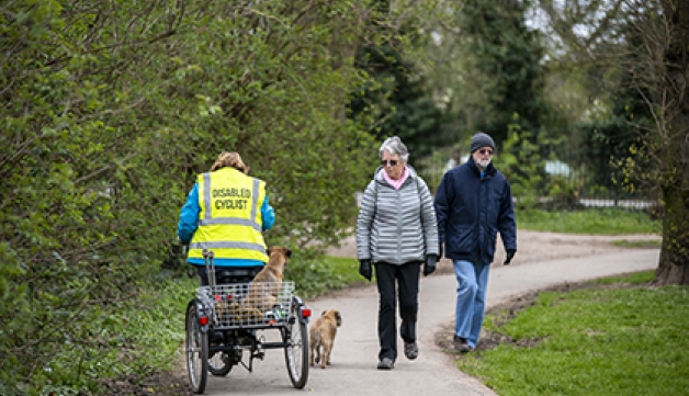 A disabled cyclist, riding a trike, rides with her down past an elderly couple on a path.